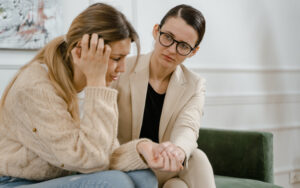Two female employees are sitting on a couch holding hands. One looks close to tears, while the other looks calm and sympathetic.